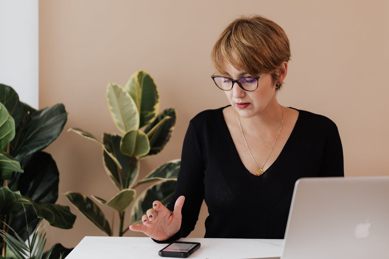 cta-01 Concentrated female manager in casual outfit and eyeglasses reading message on smartphone while sitting at table with laptop in cozy workspace