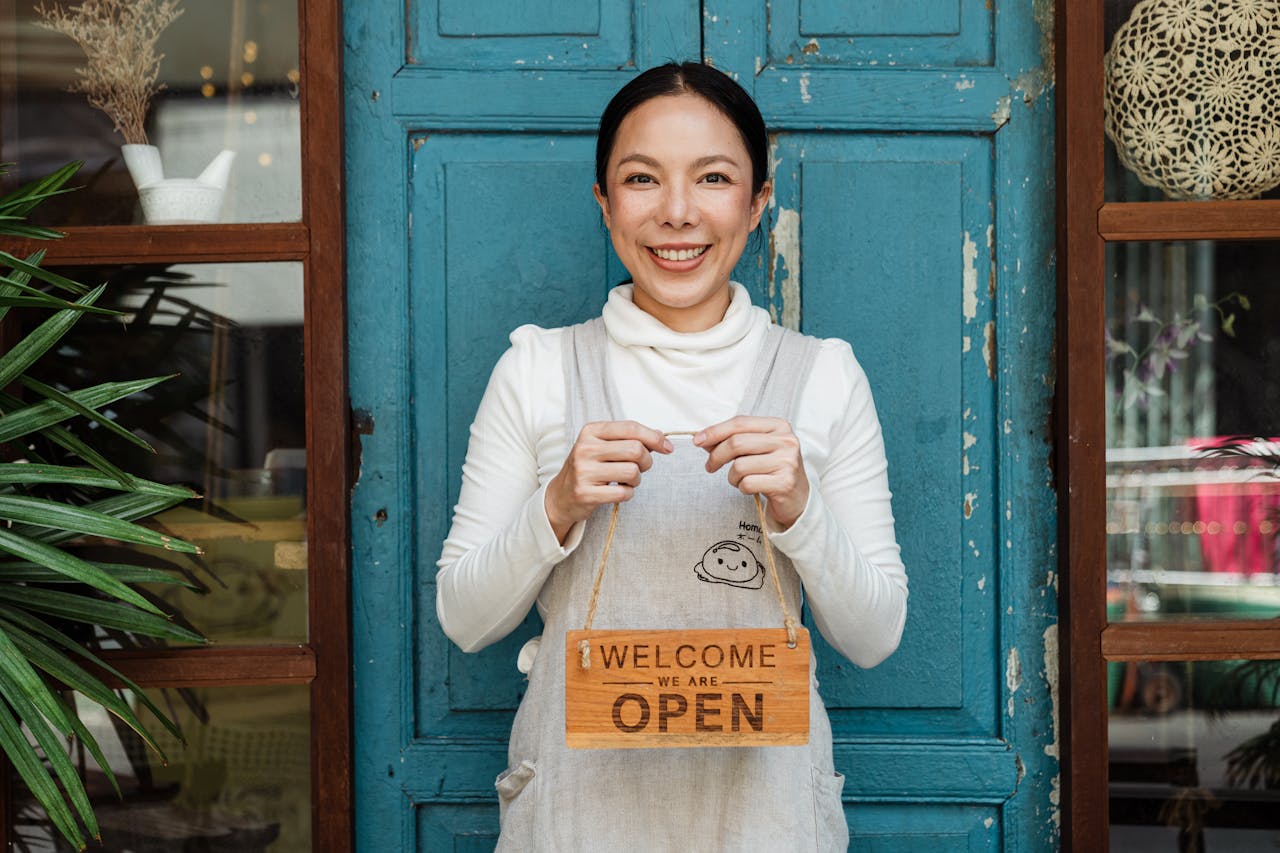 programs-03 Cheerful ethnic female cafeteria owner in apron demonstrating cardboard signboard while standing near blue shabby door and windows after starting own business and looking at camera